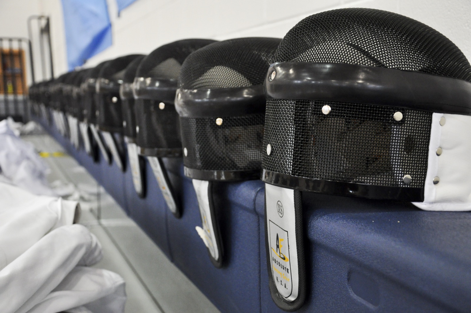 A row of fencing masks lined up on a bench beside folded white fencing jackets